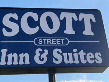 A hotel room with a bed, a desk with a chair, a small table, a trash bin, and a window with curtains. at Scott Inn Suites - Downtown Houston