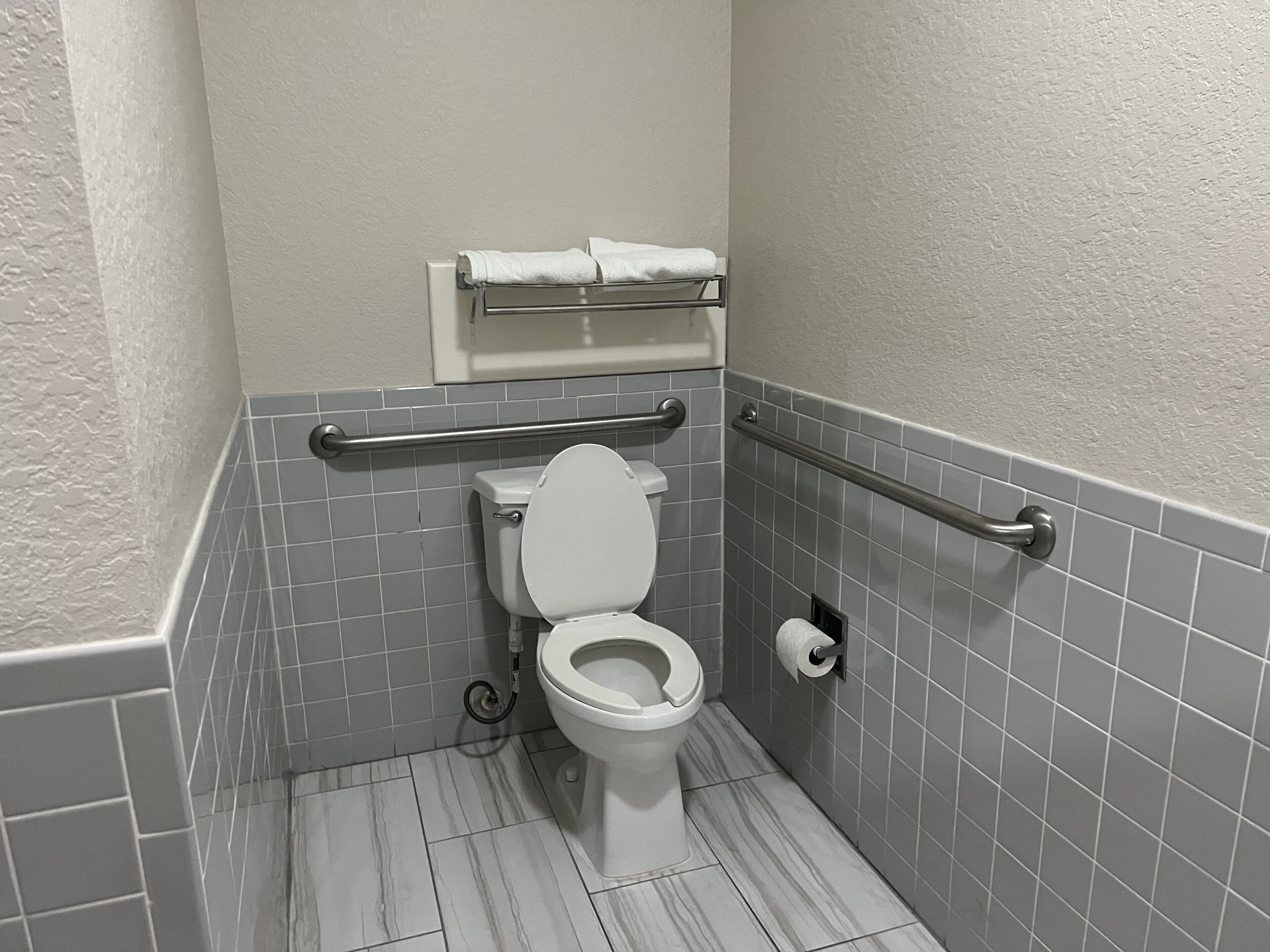 A bathroom with a white toilet, grab bars, a towel rack, and grey tiled walls and floor.