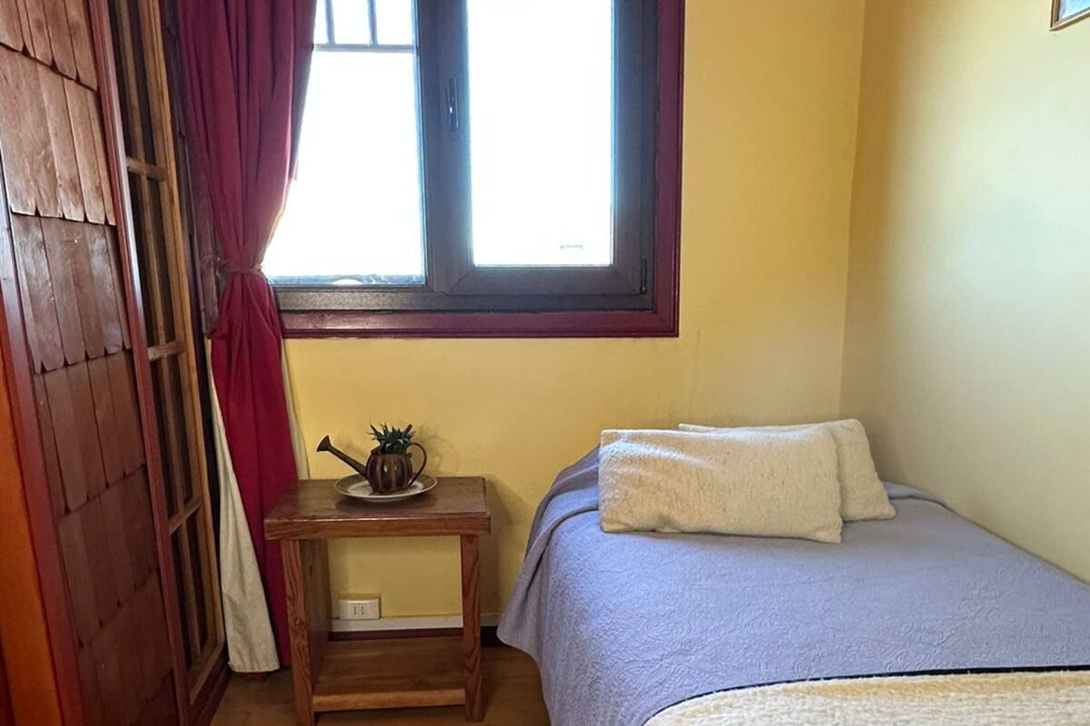 A bedroom with a wooden floor, a bed with a blue and white duvet, a small wooden nightstand with a plant, and a window with curtains.