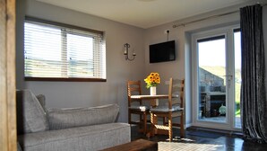 A living room with a grey sofa, wooden chairs, a table with sunflowers, a TV mounted on the wall, and a sliding glass door leading outside.