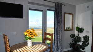 A dining area with a round wooden table, a white cushioned chair, a vase with sunflowers, a large potted plant, and a view of the outdoors through a glass door.