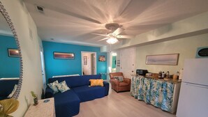A living room with a blue sofa, a brown armchair, and a kitchen area with a refrigerator and a countertop. - OBX~Beach~Sunsets~Bike Path~Mini Golf~Lighthouses~Shops~Walking Distance~Sunrise (Kill Devil Hills)