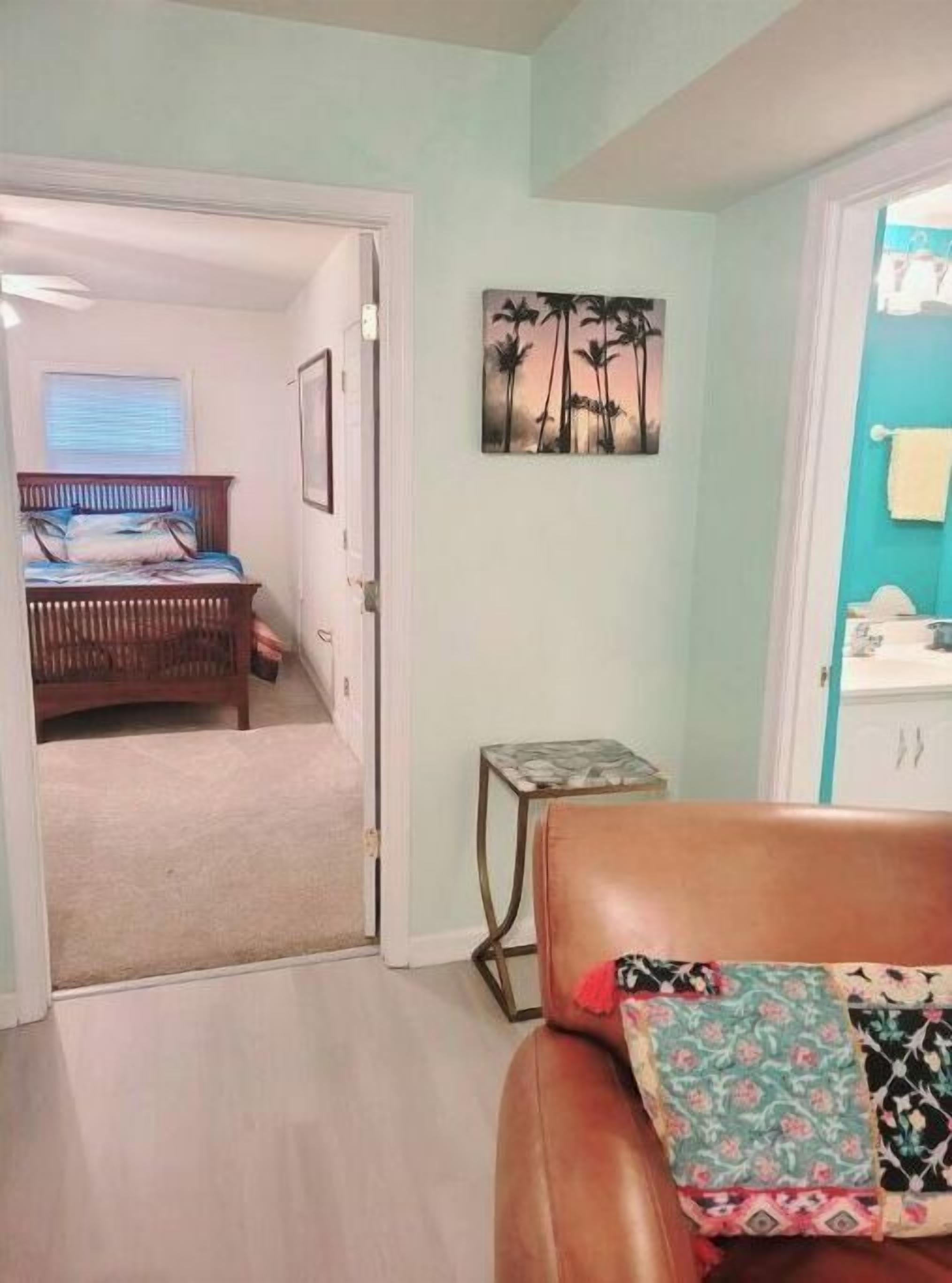 Un dormitorio con cama, una puerta que da a otra habitación, una mesita pequeña y una pared con un cuadro de motivos tropicales.