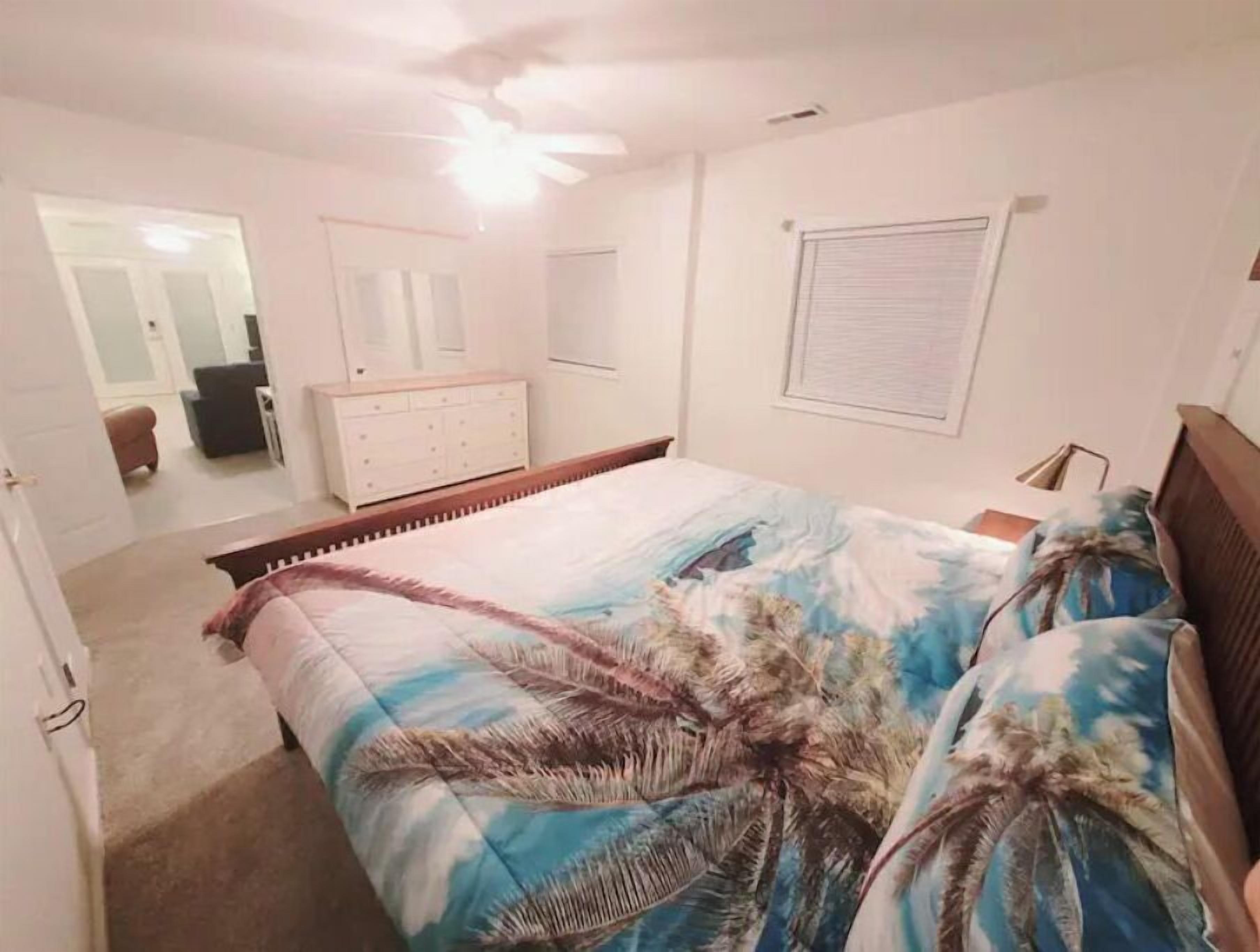 Une chambre à coucher comprenant un lit, une commode, un ventilateur de plafond et une fenêtre avec des stores.