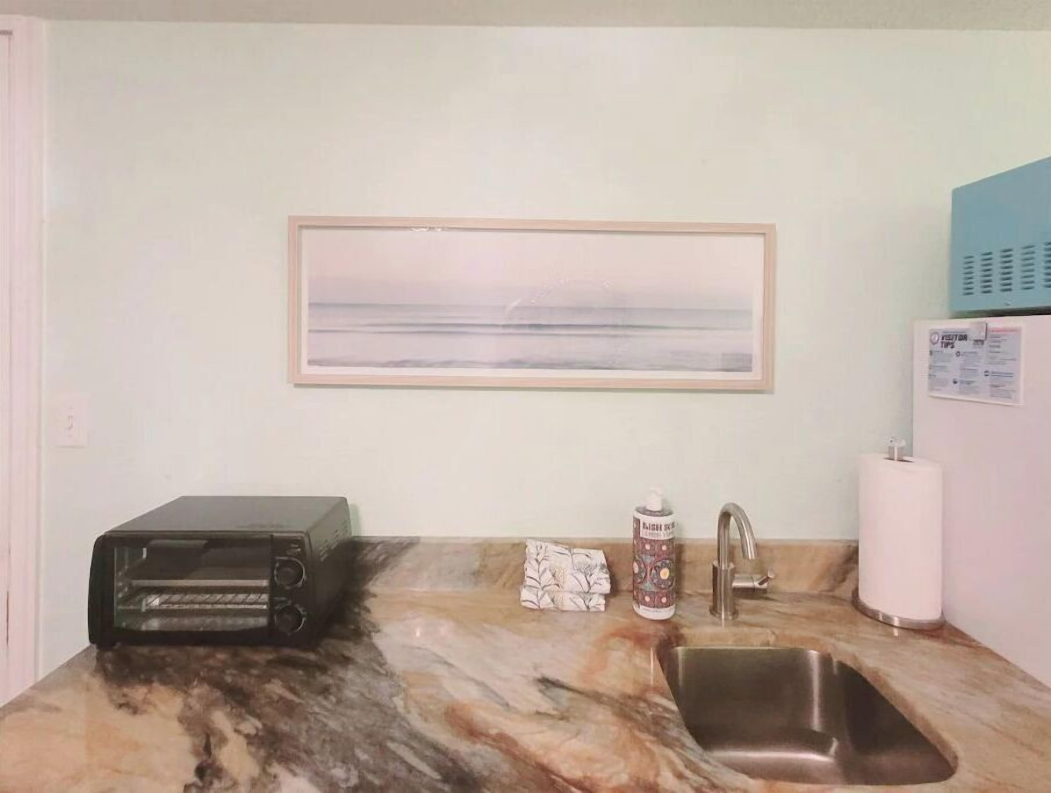 Een keuken met een marmeren aanrecht, een broodrooster, een spoelbak, een kraan, een lijst met een foto van een strand en een informatiebord voor gasten.