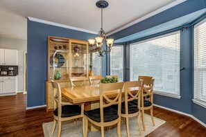 A dining area with a wooden table, four chairs, a chandelier, and a large window with blinds.