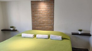 A neatly made bed with green bedding and three rolled towels, a brick accent wall, and two small potted plants on floating shelves. - My Depa Vanilla. In the heart of Cd. Valleys. (Ciudad Valles)