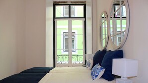 Quarto de hotel com cama, mesa de cabeceira com abajur, duas espelhos redondos na parede e varanda com vista para um edifício.