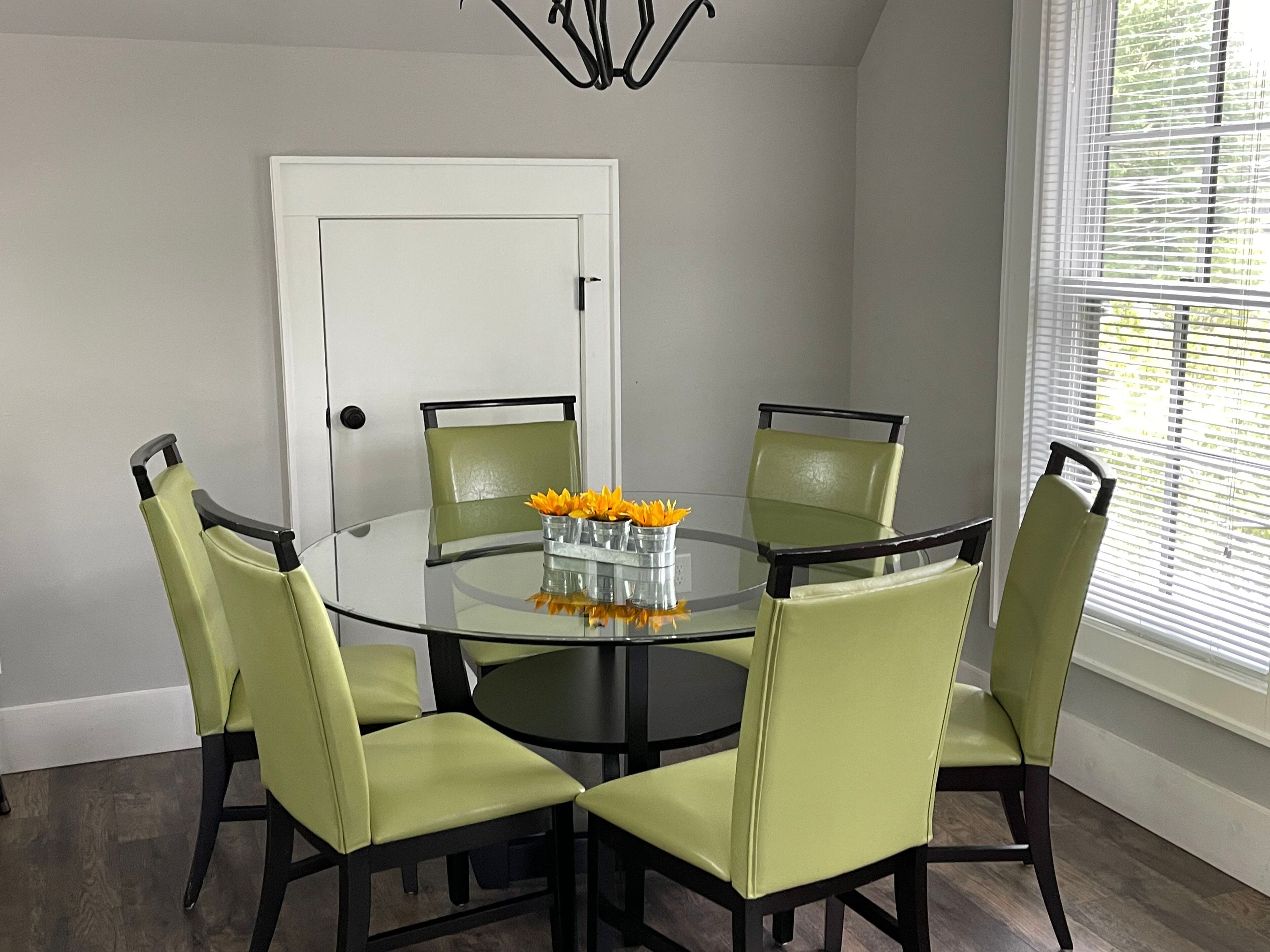 Une table ronde en verre, quatre chaises à revêtement vert, une porte blanche et une fenêtre avec des stores.