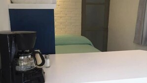 Una stanza compatta con un letto addossato a una parete in mattoni bianchi, un tavolino con una macchina da caffè e due sgabelli.