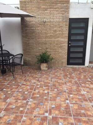 Eine Terrasse mit Fliesenboden, einer Ziegelwand, einer schwarzen Tür und einem kleinen Topfpflanzen.