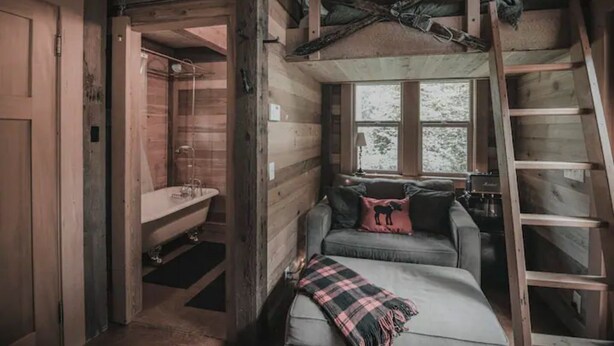Interior de una cabaña con cama en el altillo, baño peque ño y ventana con vistas a árboles.
