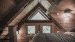 Un dormitorio con techo inclinado, vigas de madera, tragaluces, dos ventanas, una cama y una mesita de noche.