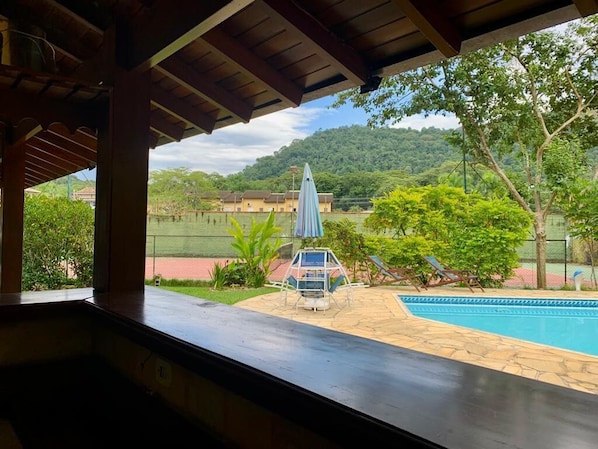 A view from a covered area overlooking a swimming pool, with a gazebo and lounge chairs in the foreground and a mountainous landscape in the background. - Pousada Brazish (Paraty)