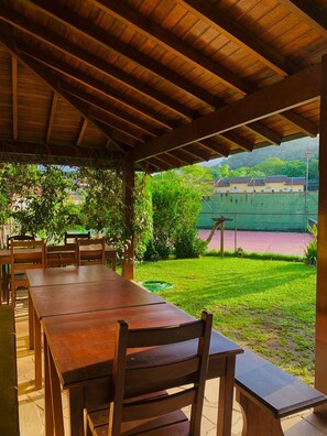 An outdoor wooden pergola with tables and chairs, overlooking a tennis court and greenery.