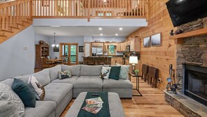A spacious living room with a large sectional sofa, a fireplace, and a wooden staircase leading to an upper level. - Cozy 5BR Cabin with 3 Fireplaces, Hot Tub, Fenced Yard (Ellijay)
