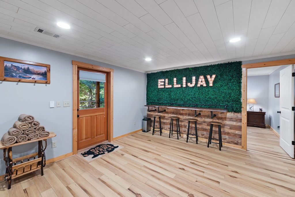 Huoneessa on puulattia, ovi, seinälle asennettu baaritiski jakkaroineen sekä vihreä seinä, johon on kirjoitettu nimi ’ELLIJAY’.