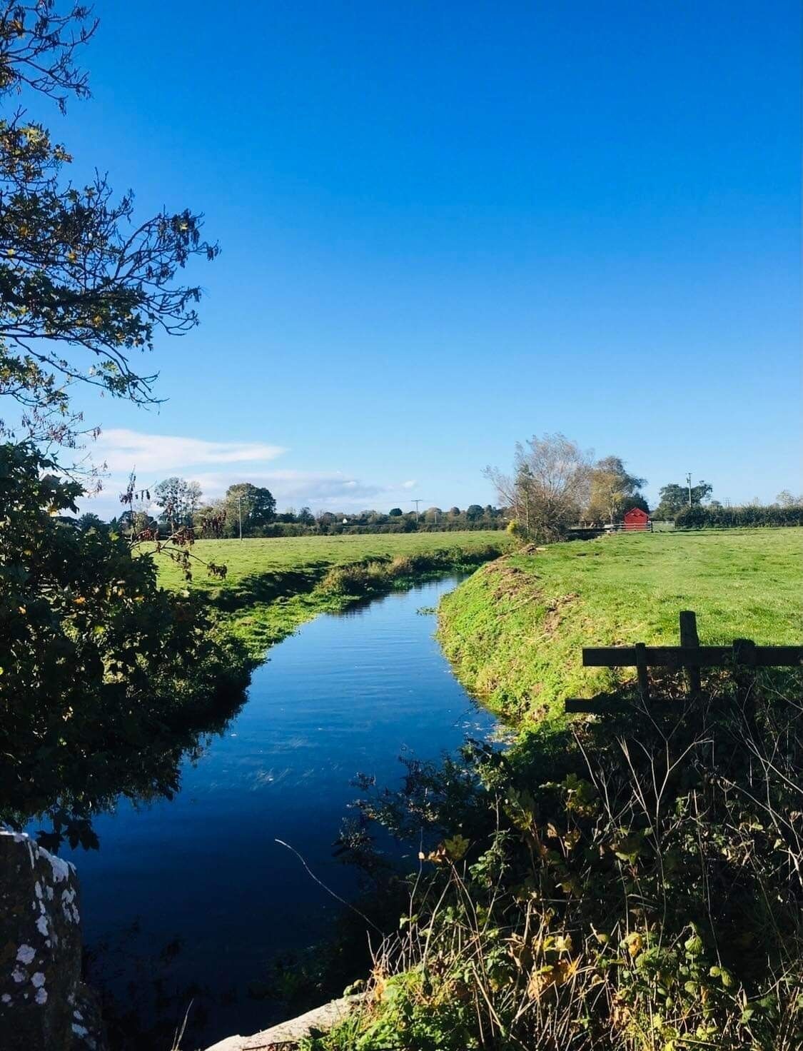 Een landelijk landschap met een smal waterkanaal, groene velden en een rode bebouwing in de verte.