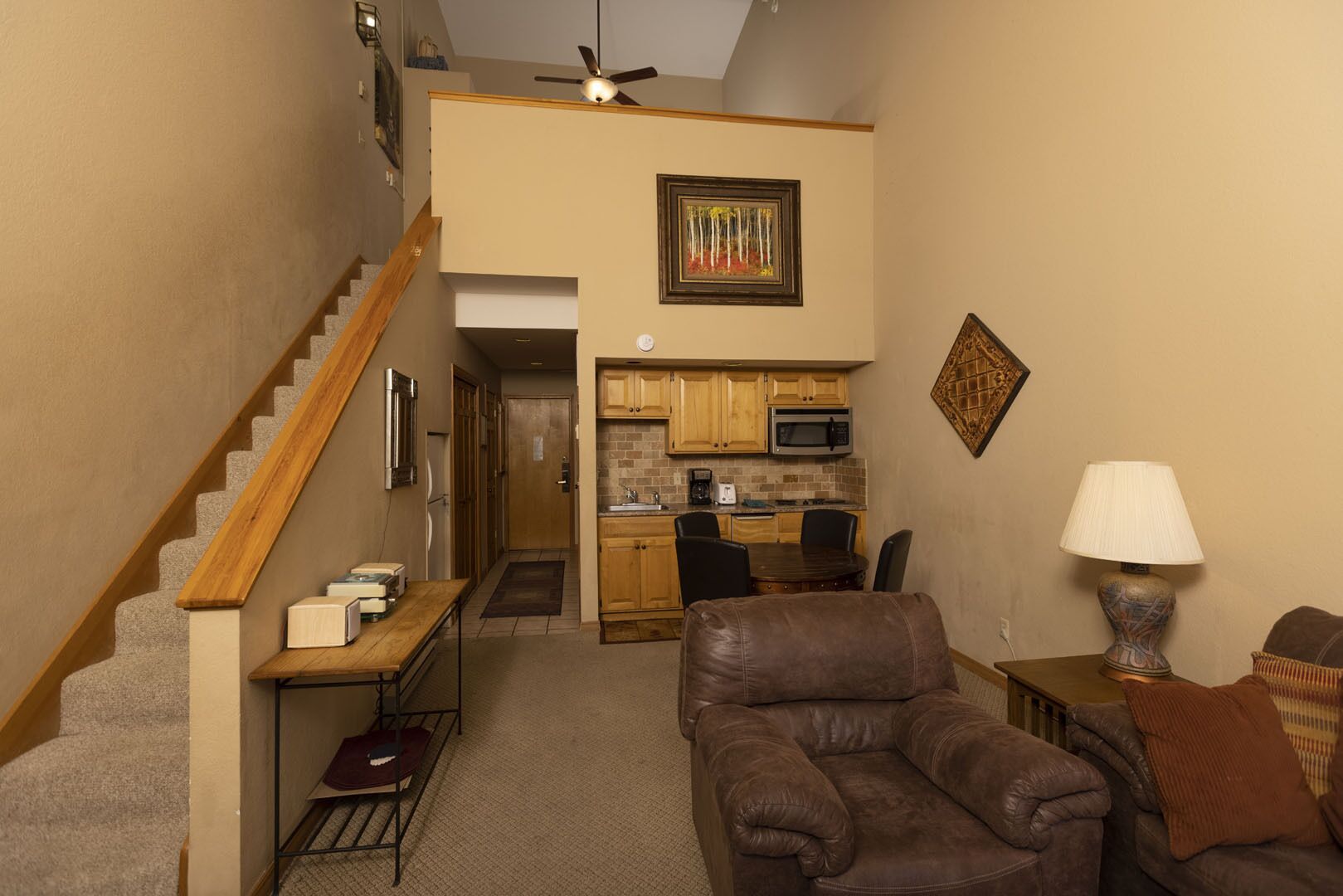 A compact living space with a staircase, a kitchen area, and a seating area with a brown leather couch and a table lamp.