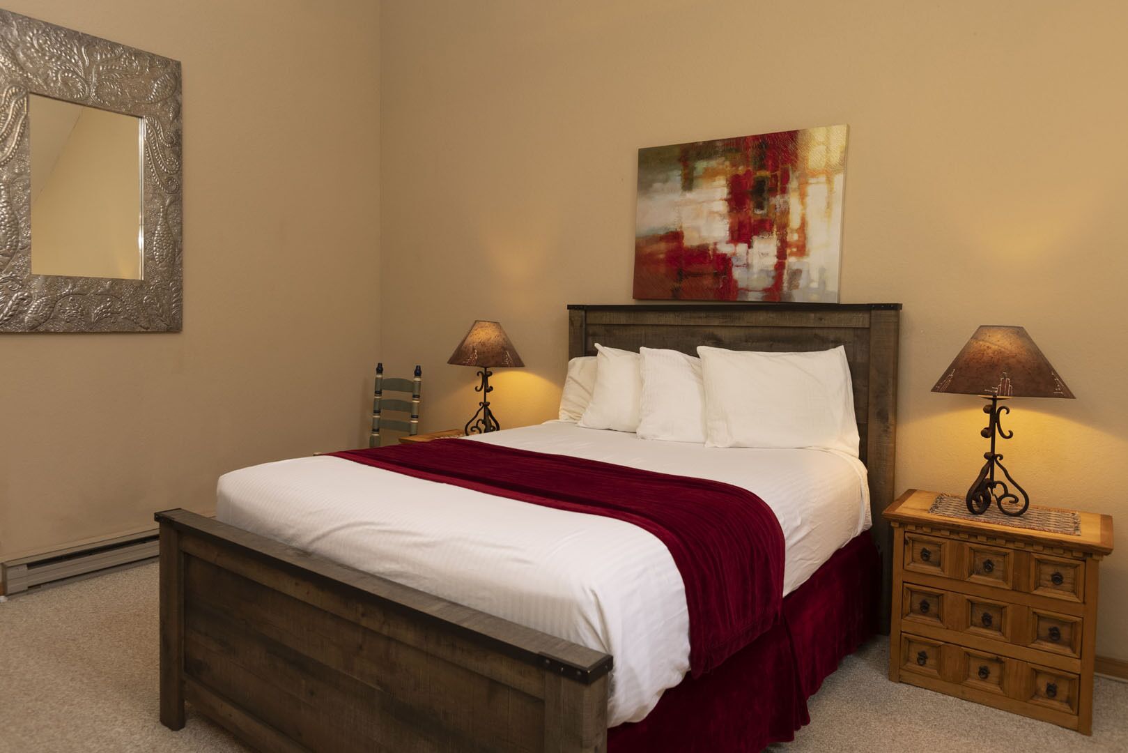 A bedroom with a wooden bed, white bedding, a red throw blanket, two bedside tables with lamps, a wooden nightstand, and a framed artwork above the bed.
