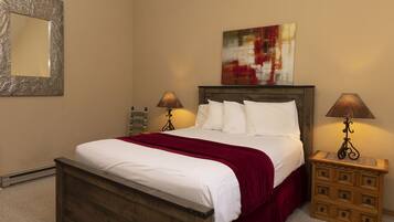 A bedroom with a wooden bed, white bedding, a red throw blanket, two bedside tables with lamps, a wooden nightstand, and a framed artwork above the bed.