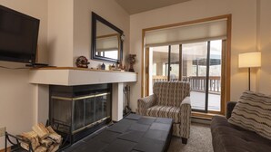 A living room with a fireplace, a television mounted above it, a patterned armchair, a sofa, and a large window with a view of the outdoors. - Cozy Studio for 2 at Purgatory - Kendall 232 (Durango)