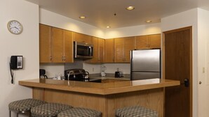 A modern kitchen with wooden cabinets, a stainless steel refrigerator, a microwave, a kettle, and a coffee maker. There are three bar stools and a phone on the wall.