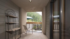 A balcony with a metal chair and a wooden table, a metal shelf unit, and a view of a forested area.