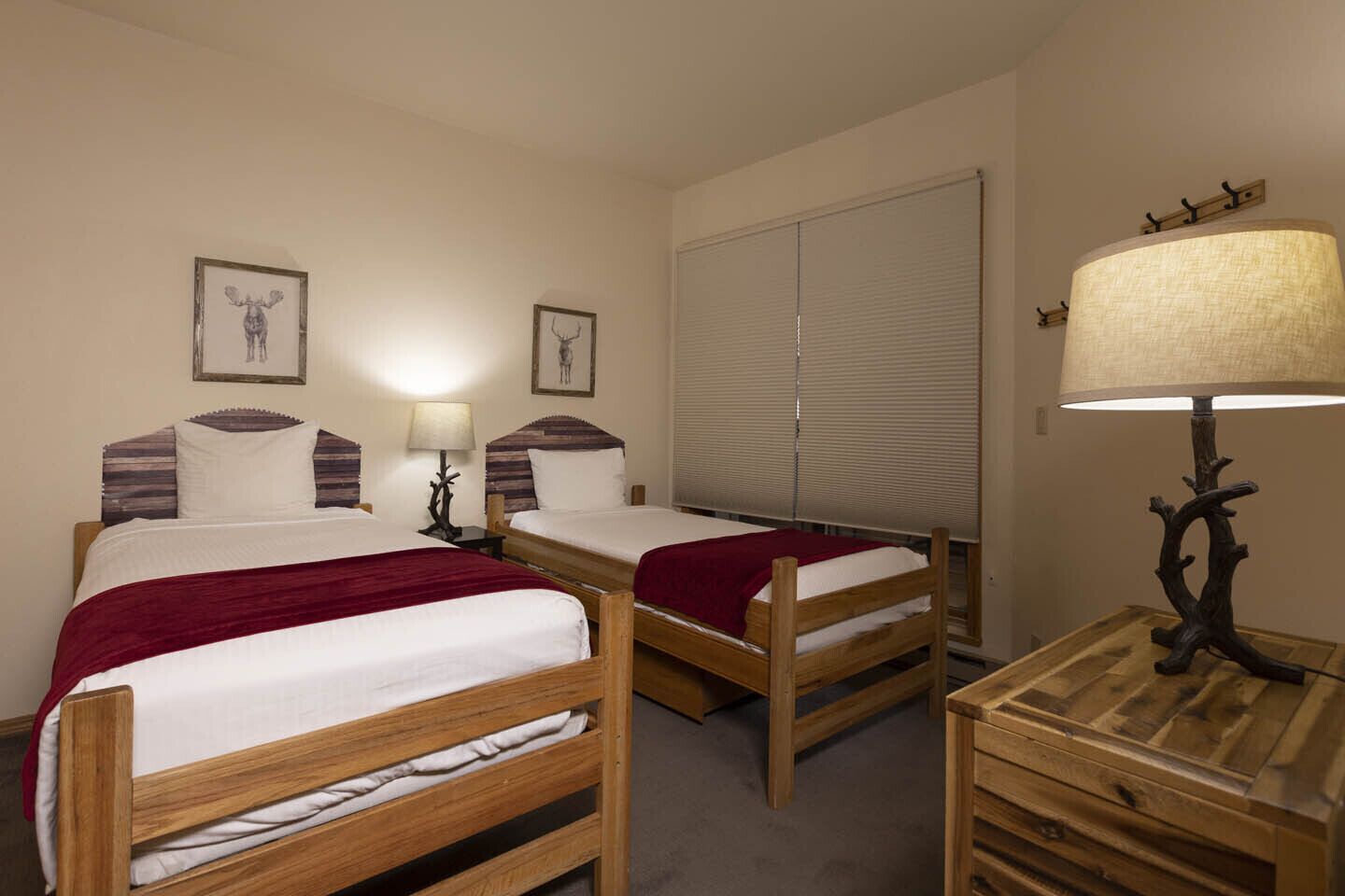 A double room with two wooden beds, each with a red blanket, a bedside table with a lamp, and a large window with blinds.