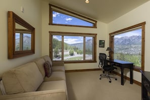 Una habitación con un sofá beige, un escritorio con una silla y ventanas grandes con vistas a la montaña.
