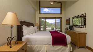Un dormitorio con una cama grande, una mesita de noche, un aparador, una lámpara y vistas a un bosque por la ventana.