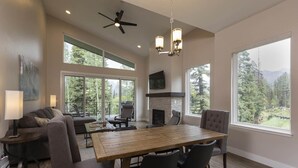 A spacious living room with a large wooden dining table, comfortable seating, a fireplace, and large windows offering views of a forested area. - Tacoma Vista 147 - Managed by Purgatory Result (Durango)