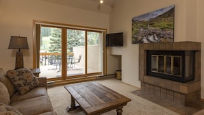 Una sala de estar con un sofá, una mesa de centro de madera, una chimenea, un televisor montado en la pared y un ventanal corredero que da a un patio exterior.
