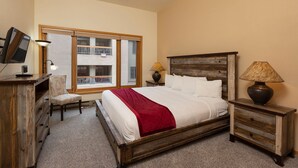 A bedroom with a wooden bed, a TV on a dresser, a chair, and a window with a view of a building. - Village Center 402 - Managed by Purgatory Resort (Durango)