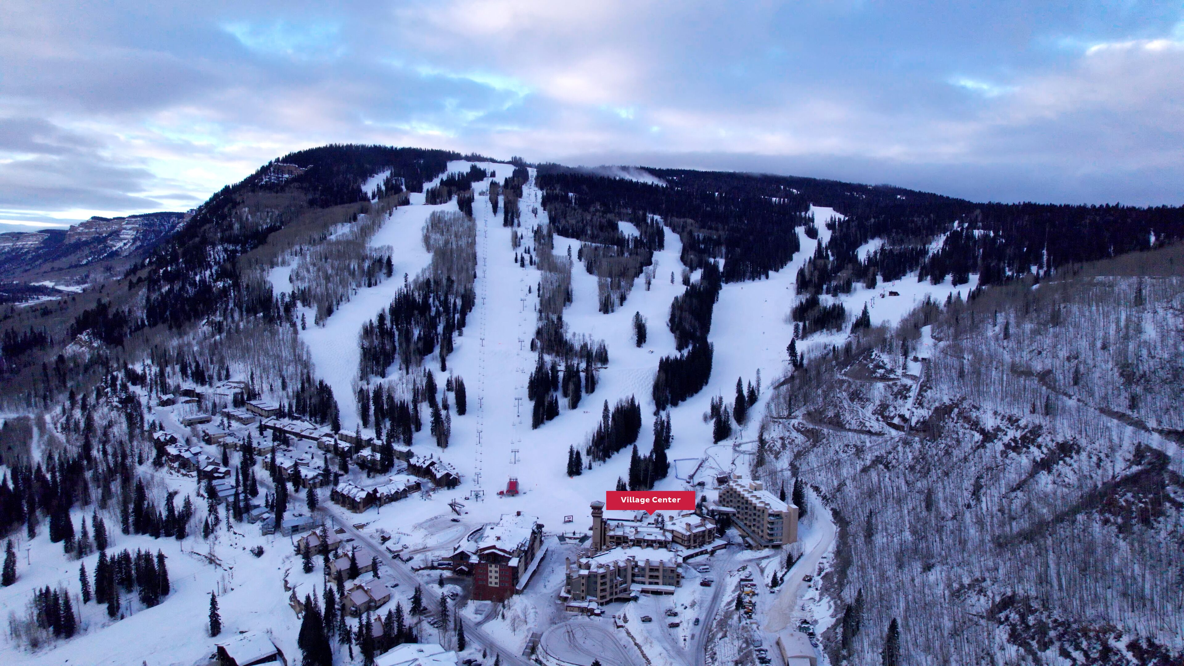 A snow-covered village center with buildings and roads, surrounded by mountains and forests.