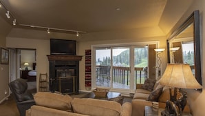 A living room with a fireplace, a sofa, a coffee table, and a lamp. There is a sliding glass door leading to a deck with outdoor furniture.