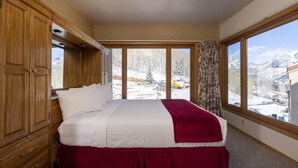 A bedroom with a large bed, wooden wardrobe, and a view of a snowy landscape through large windows. - Village Center 512 - Managed by Purgatory Resort (Durango)