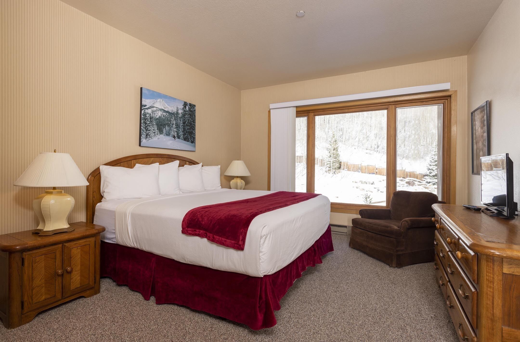 Habitación de hotel con una cama grande, dos lámparas de noche, una mesita de noche de madera, un sillón marrón, un aparador de madera y una ventana con vistas nevadas.