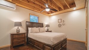 A bedroom with a wooden ceiling, a bed with a headboard, two bedside tables with lamps, a wall clock, and a window with blinds. - Cabin C at Safe Harbor Brady Mountain on Lake Ouachita! (Royal)