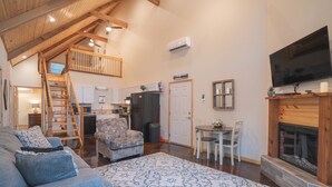 A spacious living area with a high ceiling, a fireplace, a dining table with chairs, a sofa, and a kitchen area with a refrigerator and microwave. - Cabin C at Safe Harbor Brady Mountain on Lake Ouachita! (Royal)