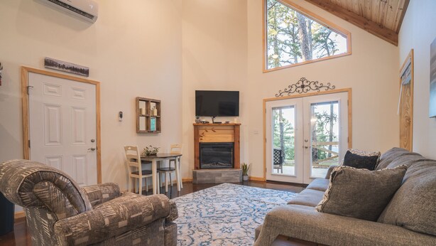 A living room with a fireplace, a television, a dining area, and a large window with a view of trees. - Cabin C at Safe Harbor Brady Mountain on Lake Ouachita! (Royal)