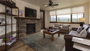 Una sala de estar con chimenea de piedra, televisor montado encima y ventilador de techo, con un amplio ventanal que ofrece vistas al exterior.