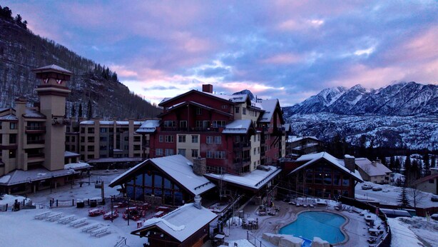Un complejo turístico cubierto de nieve con varios edificios, una zona de piscina y montañas al fondo.