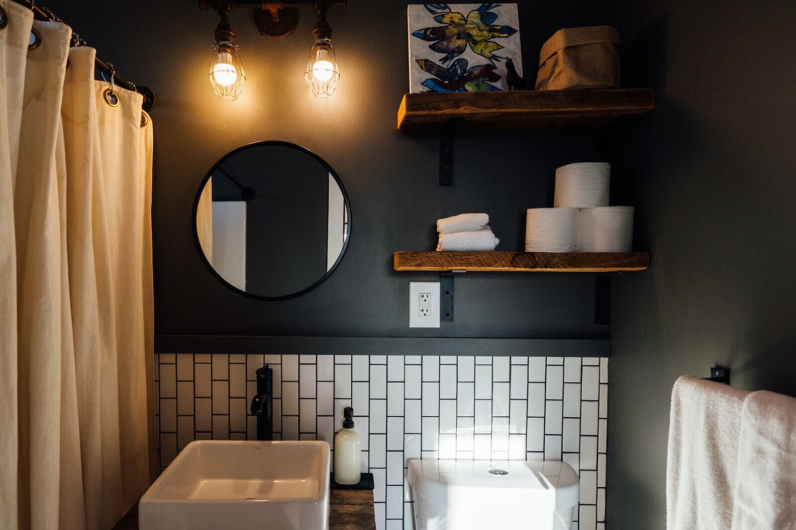 A bathroom with a white sink, a round mirror, and a toilet. There are towels and toilet paper on a shelf, and a shower curtain is partially visible.