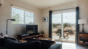 A living room with a large TV, a sofa, a lamp, and a sliding glass door leading to an outdoor patio. - Secluded Retreat-Hot Tub, Fire Pit, Outdoor Shower (Joshua Tree)