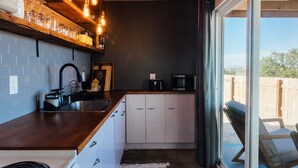 A compact kitchen with a wooden countertop, white cabinets, a black faucet, and a coffee maker. There is a window with a view of a wooden fence and trees. - Secluded Retreat-Hot Tub, Fire Pit, Outdoor Shower (Joshua Tree)
