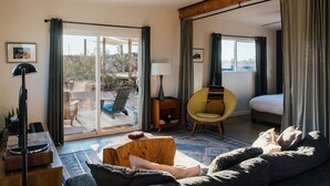 A modern hotel room with a large sliding glass door leading to a patio, a bed, a sofa, a chair, and a small table with a lamp. - Secluded Retreat-Hot Tub, Fire Pit, Outdoor Shower (Joshua Tree)