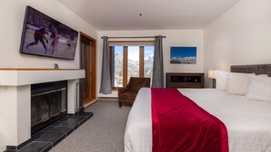 Een hotelkamer met een groot bed, een openhaard, een aan de muur bevestigde televisie, een stoel, een raam met gordijnen en een schilderij.