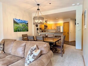 A living room with a sofa, a dining table, and chairs. There is a kitchen in the background with cabinets and appliances.