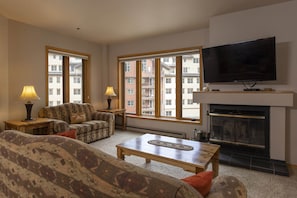 Ein Wohnzimmer mit Sofa, Sesseln und Couchtisch, einem Kamin, einem Fernseher und großen Fenstern mit Blick auf die Stadt.